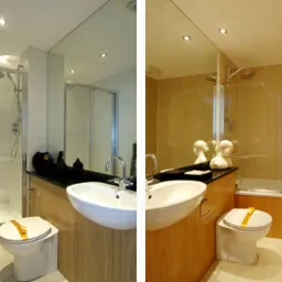 Nowoczesna łazienka z kabiną prysznicową, umywalką wpuszczoną w drewnopodobny blat i toaletą z żółtą taśmą zabezpieczającą, odbicie w lustrze ukazuje wannę z parawanem.